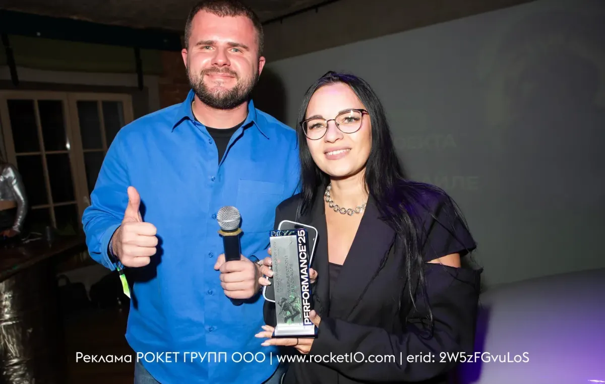 Two people at a promotional event posing with a microphone and an award Two people at a promotional event posing with a microphone and an award