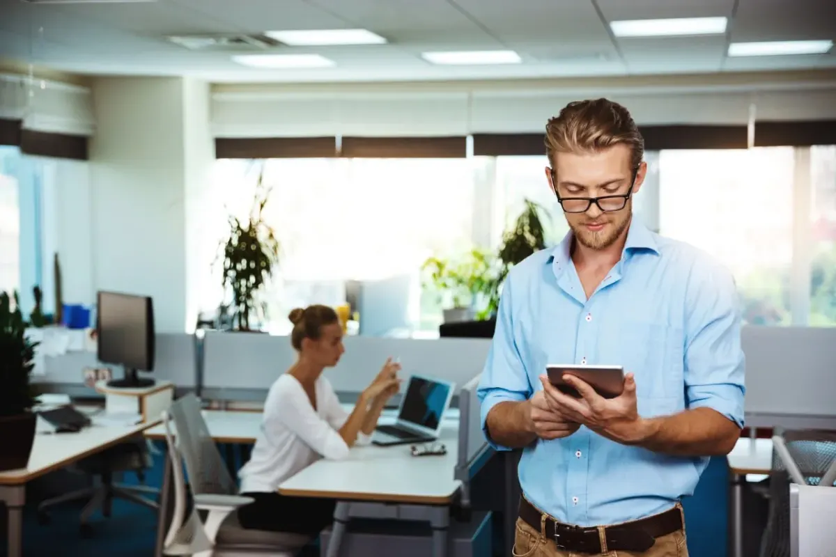 Man checking his smartphone in a modern office while a colleague works on a laptop in the background Man checking his smartphone in a modern office while a colleague works on a laptop in the background